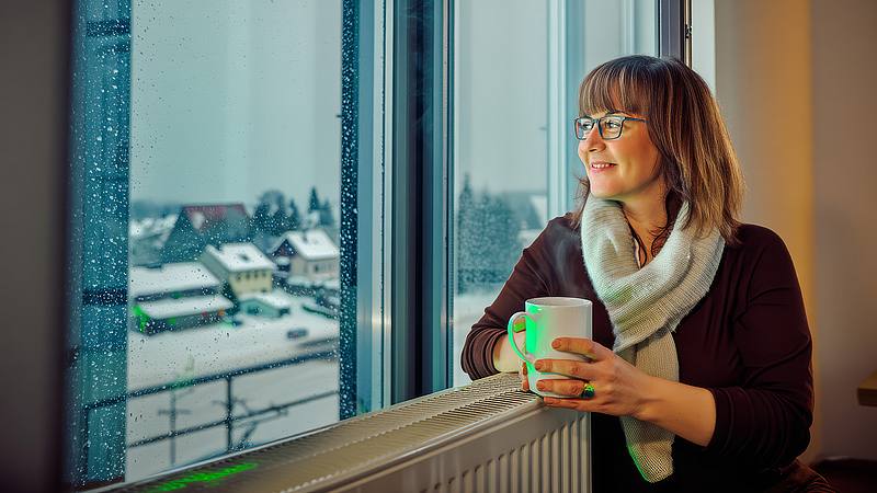 Frau schaut aus dem Fenster in eine Winterlandschaft und wärmt sich an der Heizung und einer Tasse Tee